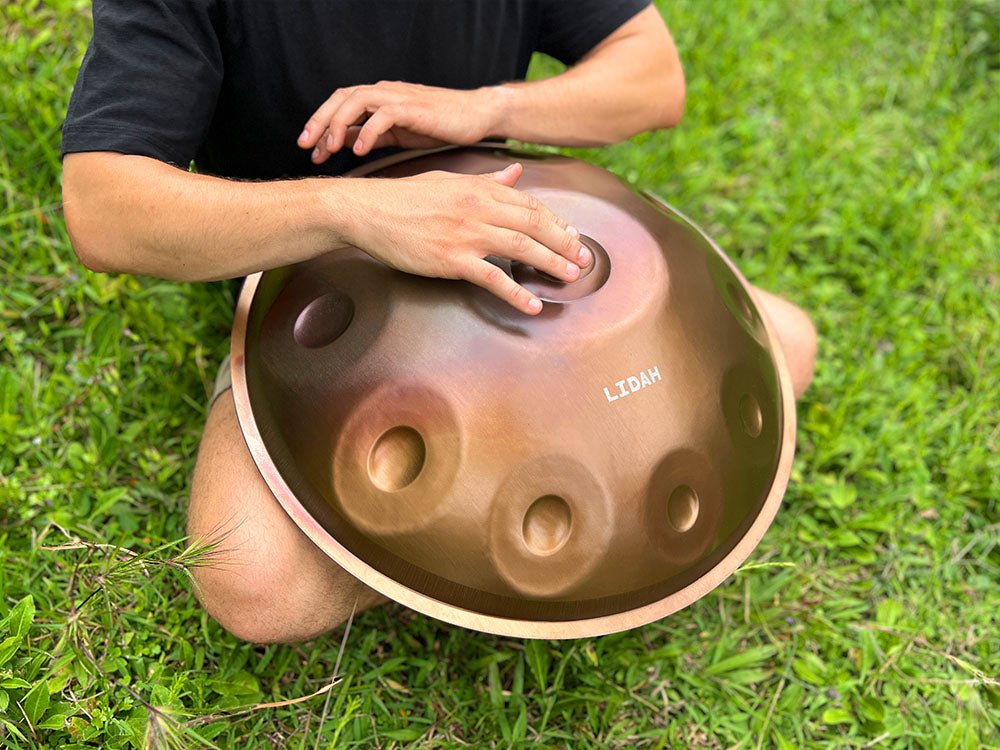 Hand playing a LIDAH handpan resting on green grass, seen from above